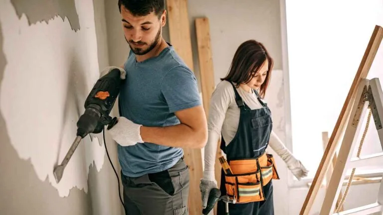 Um homem e uma mulher trabalham juntos em obra de apartamento. O homem, vestindo camiseta azul e luvas, utiliza uma furadeira para remover parte do revestimento da parede. A mulher, com macacão de trabalho e cinto de ferramentas laranja, segura uma furadeira, preparando-se para a próxima etapa.