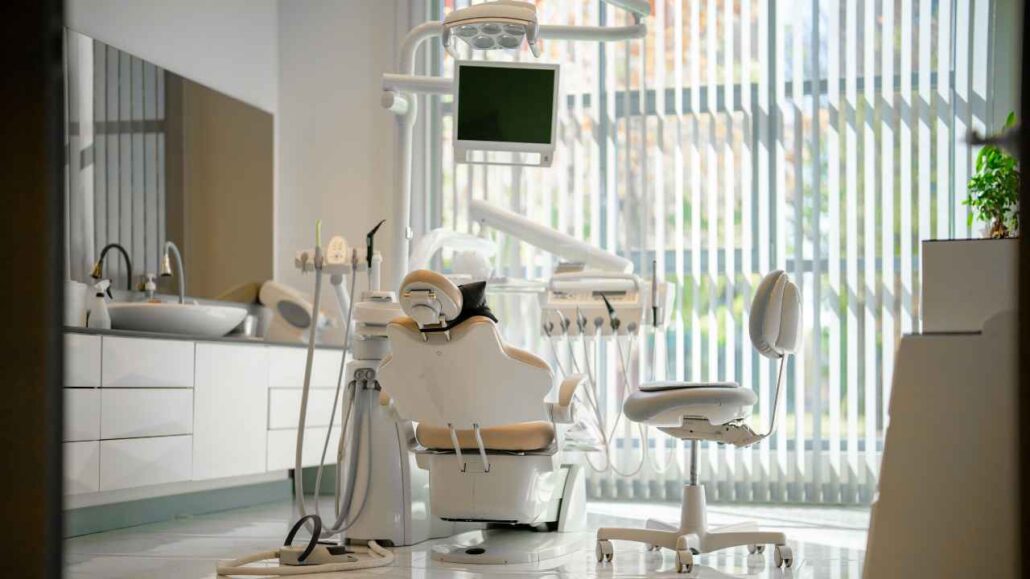 Consultório odontológico moderno com cadeira de dentista, equipamentos especializados e iluminação natural entrando pelas persianas da janela.