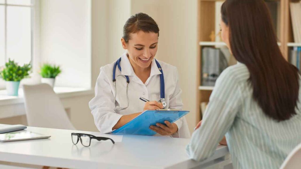 Médica sorridente faz anotações em uma prancheta enquanto conversa com uma paciente em consultório
