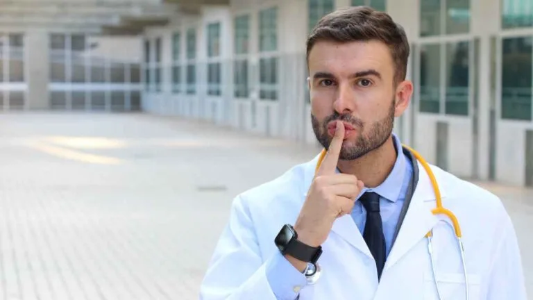 Médico de jaleco branco, com estetoscópio amarelo no pescoço, faz gesto de silêncio levando o dedo indicador aos lábios