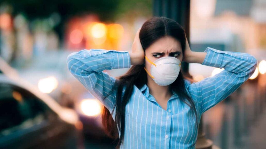 Mulher jovem usando máscara protetora cobre os ouvidos com as mãos em meio ao trânsito movimentado, mostrando incômodo com o barulho intenso da rua.