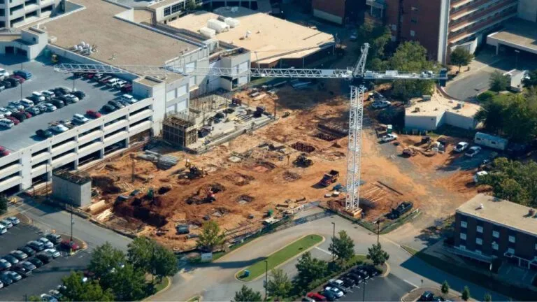 Vista aérea de uma grande obra em andamento ao lado de um hospital. A imagem mostra máquinas, guindaste e estruturas sendo erguidas.