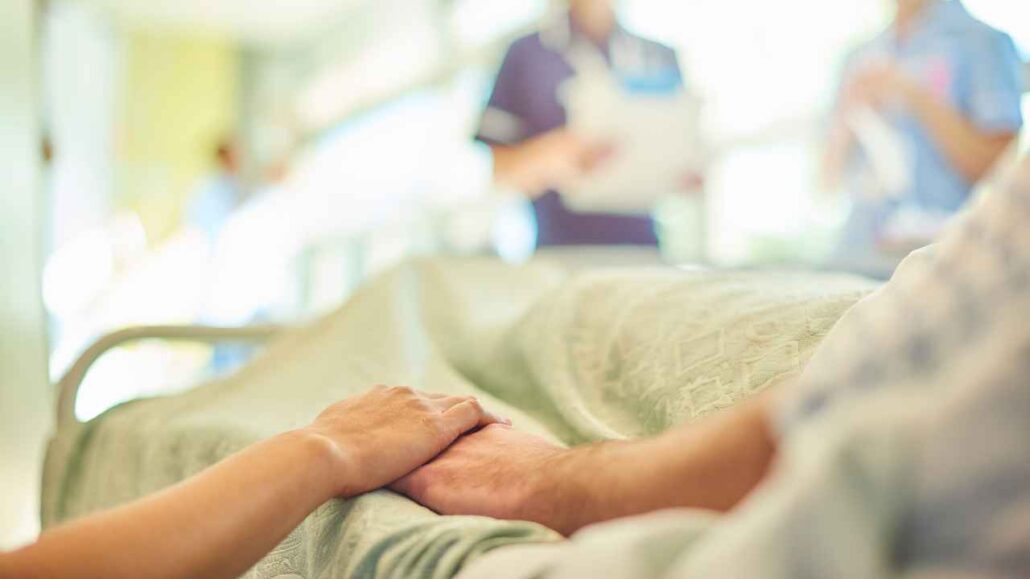 Mão de uma pessoa segurando a de um paciente deitado em um leito de consultório, transmitindo cuidado e apoio, com profissionais de saúde desfocados ao fundo.