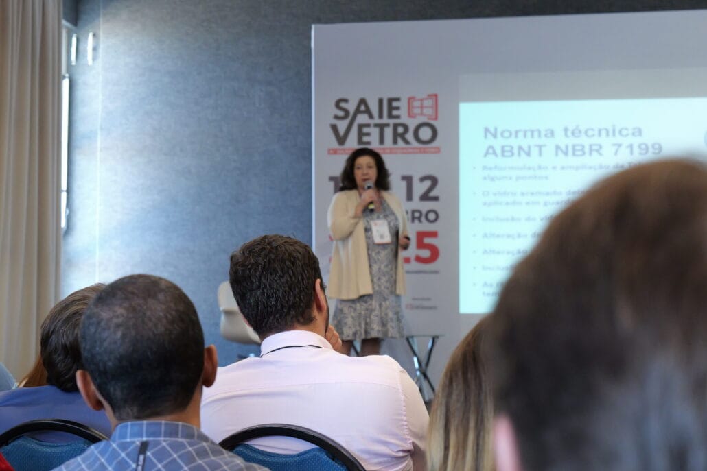 Clélia Bassetto, analista de normalização, apresenta palestra técnica durante o 18° VidroSom. Ela está no palco com microfone em mãos, ao lado de um slide projetando informações sobre a norma ABNT NBR 7199