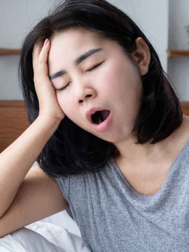 Mulher sentada na cama bocejando e apoiando a cabeça na mão, com expressão de cansaço, representando o desconforto de quem acorda exausta após uma noite mal dormida devido ao barulho noturno