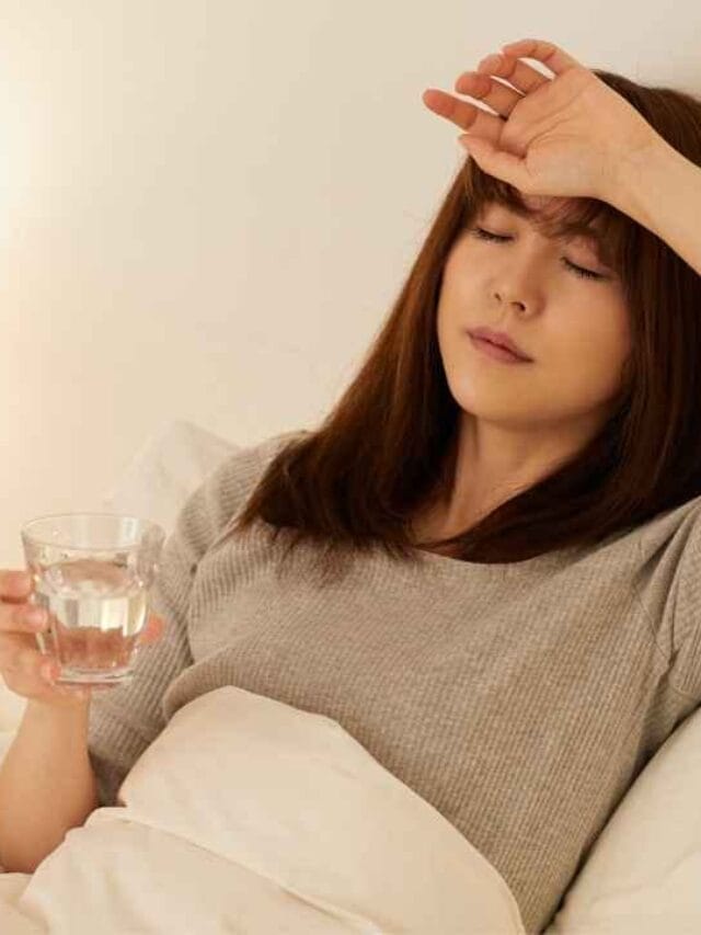 Mulher deitada na cama com expressão de cansaço, segurando um copo de água e tocando a testa, em um ambiente iluminado por abajur, representando os efeitos de imunidade baixa e fadiga.