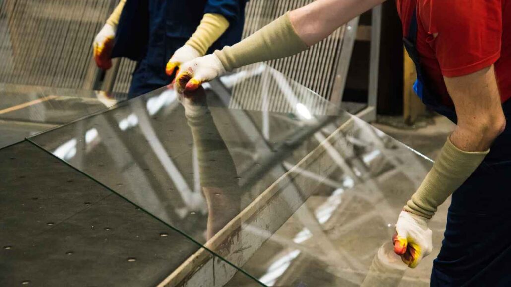 Trabalhadores com luvas de proteção manuseiam uma grande chapa de vidro em ambiente industrial.