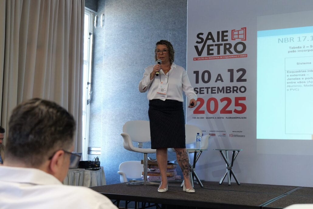 A engenheira Michele Gleice apresenta palestra no evento 18° Vidrosom. Ela está no palco segurando um microfone, com projeção de slide técnico sobre normas NBR ao fundo e o público acompanhando a apresentação.