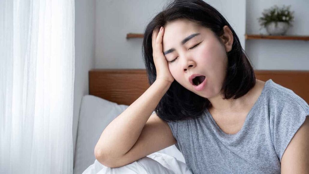 Mulher sentada na cama bocejando e apoiando a cabeça na mão, com expressão de cansaço, representando o desconforto de quem acorda exausta após uma noite mal dormida devido ao barulho noturno