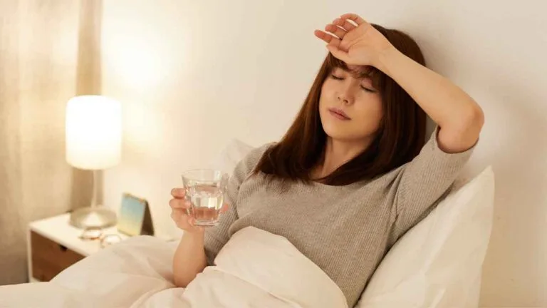 Mulher deitada na cama com expressão de cansaço, segurando um copo de água e tocando a testa, em um ambiente iluminado por abajur, representando os efeitos de imunidade baixa e fadiga.