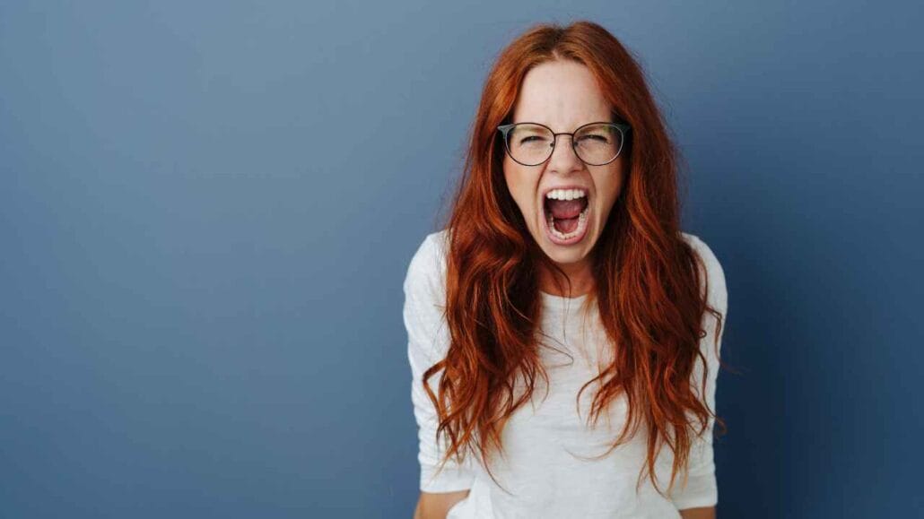 Mulher ruiva usando óculos e camiseta branca grita com expressão de irritação, em frente a uma parede azul, representando incômodo e estresse causados por barulhos excessivos no ambiente.