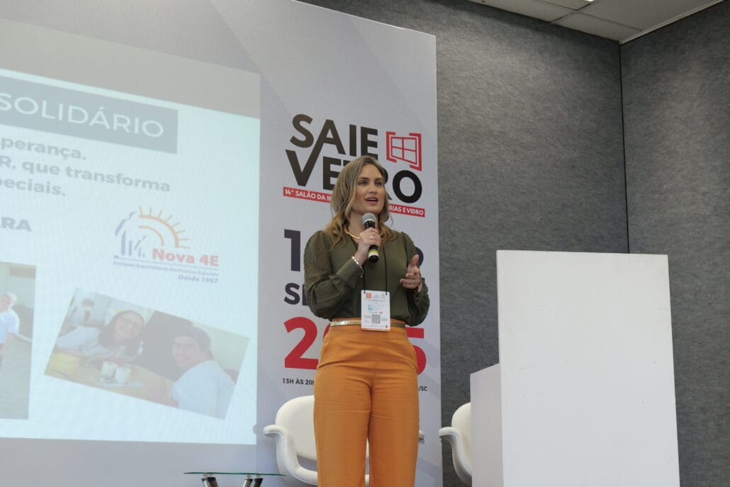 Nicole, representante da Atenua Som, apresenta palestra no 18° VidroSom. Ela está em pé no palco com microfone em mãos, ao lado de uma tela projetando informações sobre tendências globais em acústica.