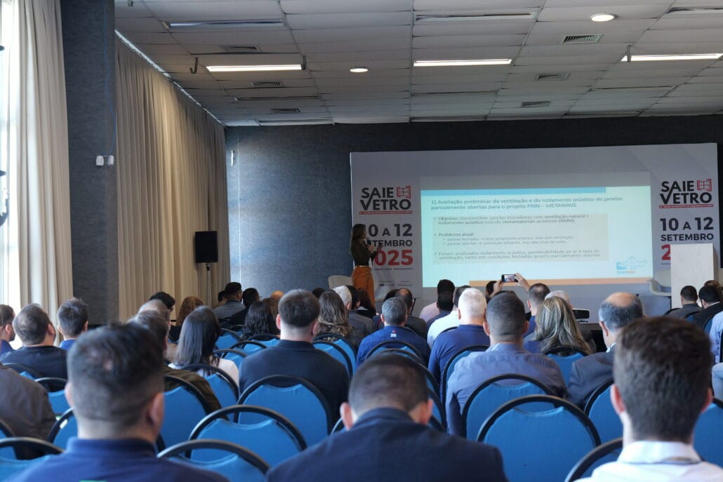 Apresentação de Nicole representando a AtenuaSom durante palestra no evento VidroSom 2025, com plateia assistindo e slide projetado