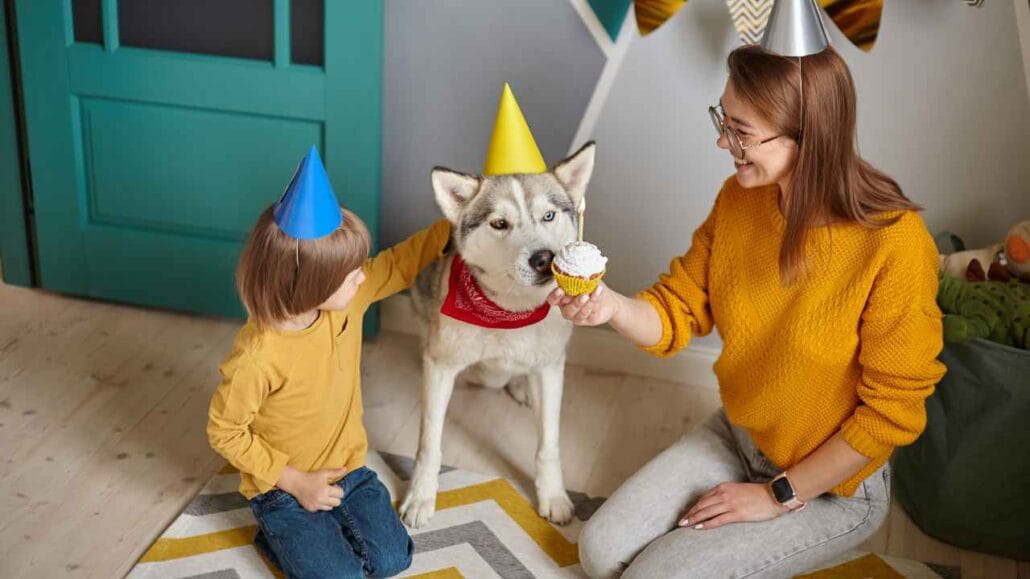 Cachorro usando chapéu de festa enquanto uma criança e uma mulher oferecem um cupcake