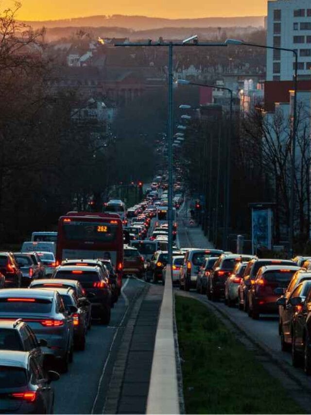 Rua urbana totalmente congestionada ao entardecer, com longas filas de carros parados nos dois sentidos, luzes de freio acesas e edifícios ao fundo, iluminados pela luz do pôr do sol.