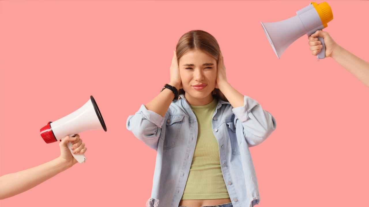 Jovem tampando os ouvidos enquanto duas pessoas seguram megafones direcionados a ela. Fundo rosa sólido