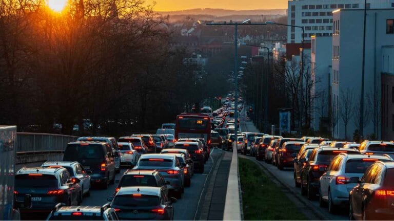 Rua urbana totalmente congestionada ao entardecer, com longas filas de carros parados nos dois sentidos, luzes de freio acesas e edifícios ao fundo, iluminados pela luz do pôr do sol.
