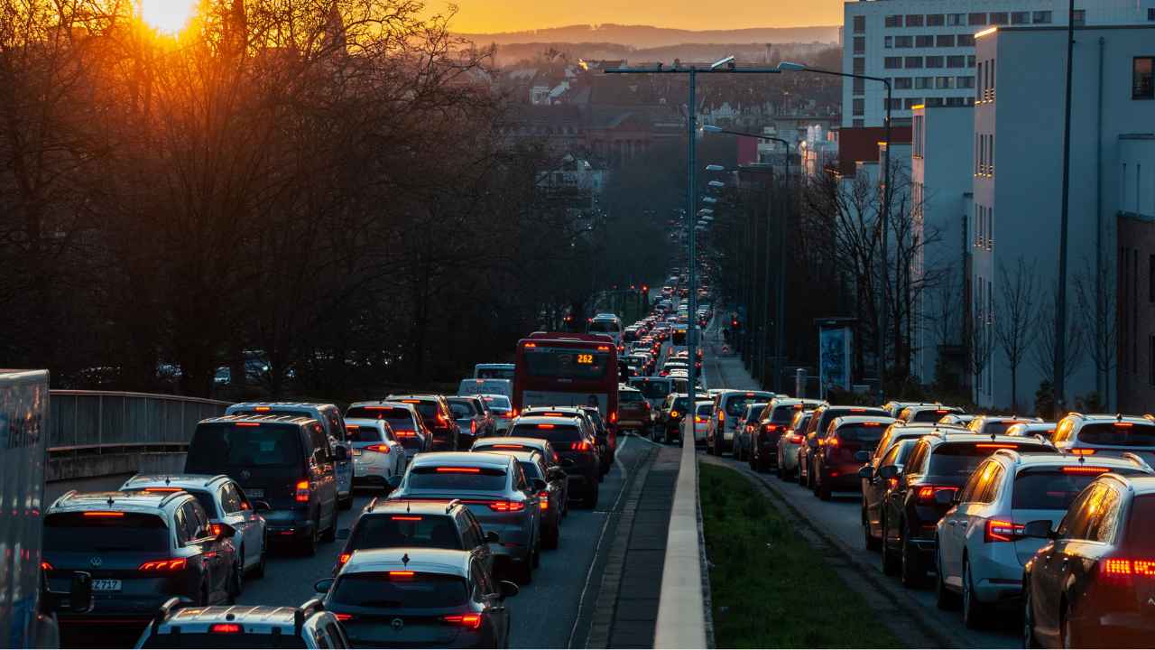 Rua urbana totalmente congestionada ao entardecer, com longas filas de carros parados nos dois sentidos, luzes de freio acesas e edifícios ao fundo, iluminados pela luz do pôr do sol.