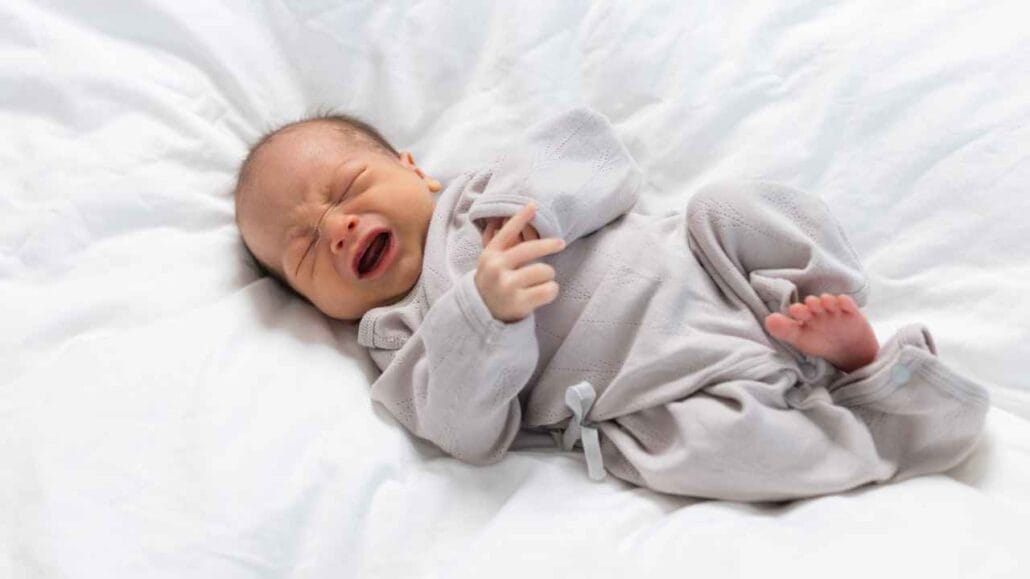 Bebê deitado em cama branca chorando, demonstrando desconforto ou irritação por não conseguir dormir
