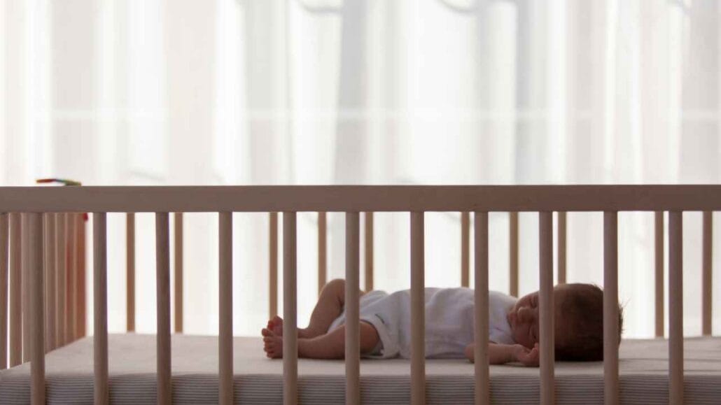 Bebê dormindo de lado em berço branco, em quarto iluminado por luz natural. De frente a uma janela antirruido.