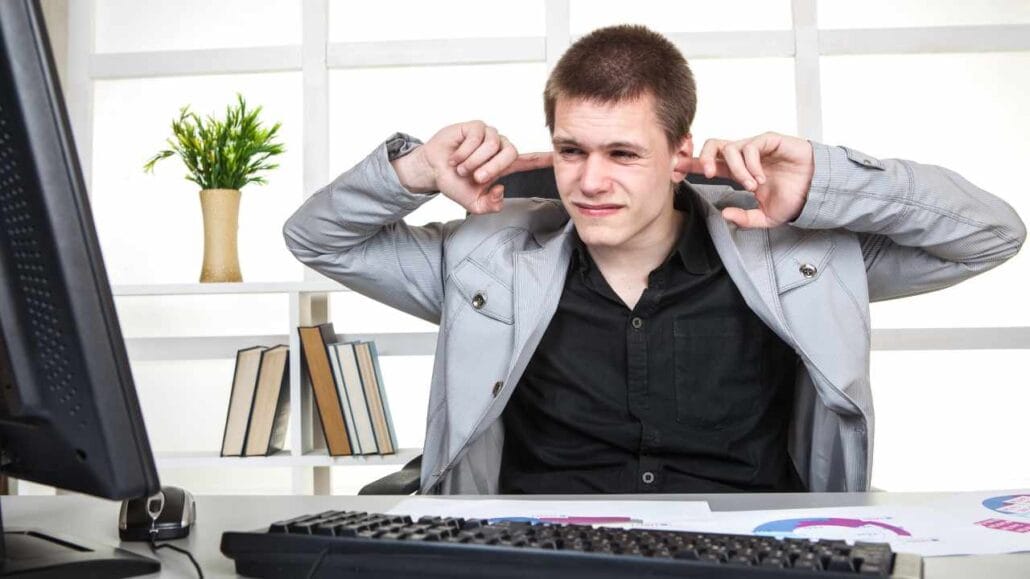 Homem sentado em frente ao computador com expressão de desconforto, tampando os ouvidos para se proteger do excesso de barulho no escritório.
