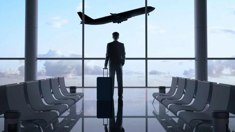 Homem de terno em um aeroporto, segurando uma mala enquanto observa um avião decolando pela janela