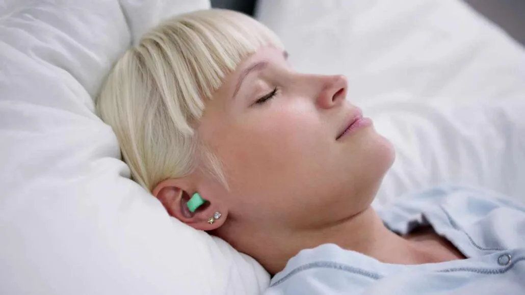 Mulher dormindo de lado em uma cama branca, usando protetor auricular verde para reduzir ruídos