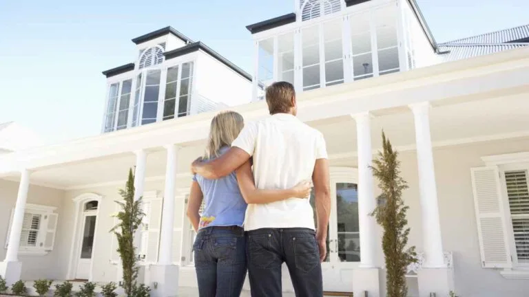 Casal em frente de uma casa branca grande, a qual possui janelas e portas antirruído. Eles observam a casa e admiram as soluções acústicas implementadas.