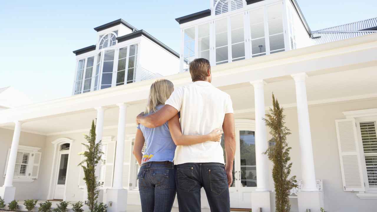 Casal em frente de uma casa branca grande, a qual possui janelas e portas antirruído. Eles observam a casa e admiram as soluções acústicas implementadas.