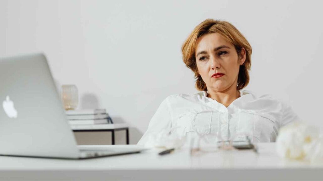 Mulher sentada à mesa olhando para um laptop com expressão séria e insatisfeita, possivelmente devido a desconcentração