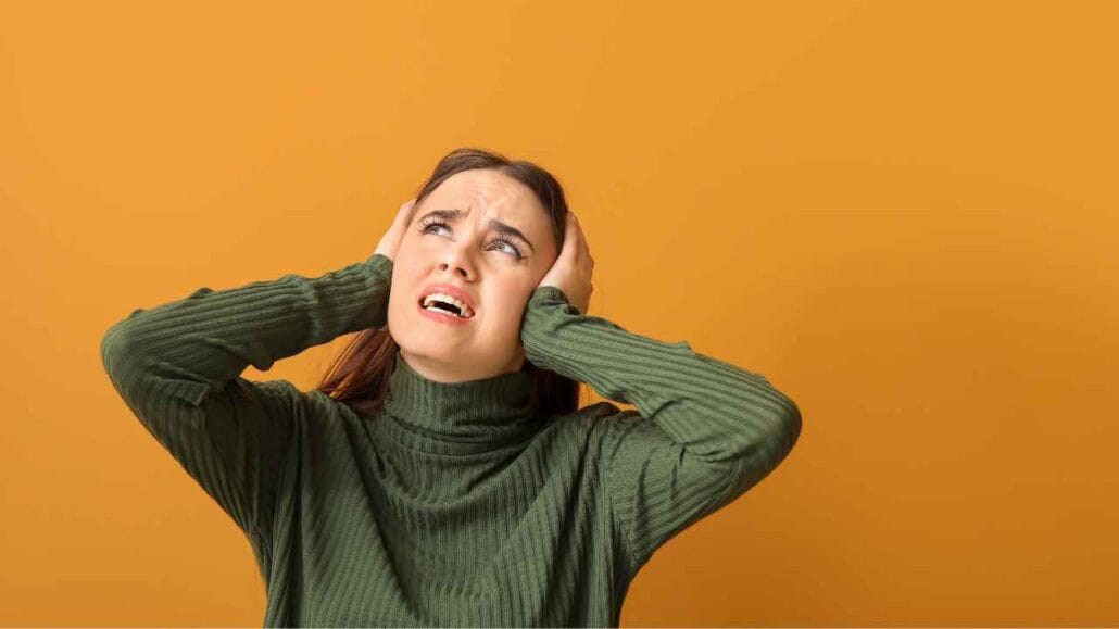Mulher de blusa verde em frente a fundo laranja, segurando a cabeça com as mãos e demonstrando desconforto devido ao barulho