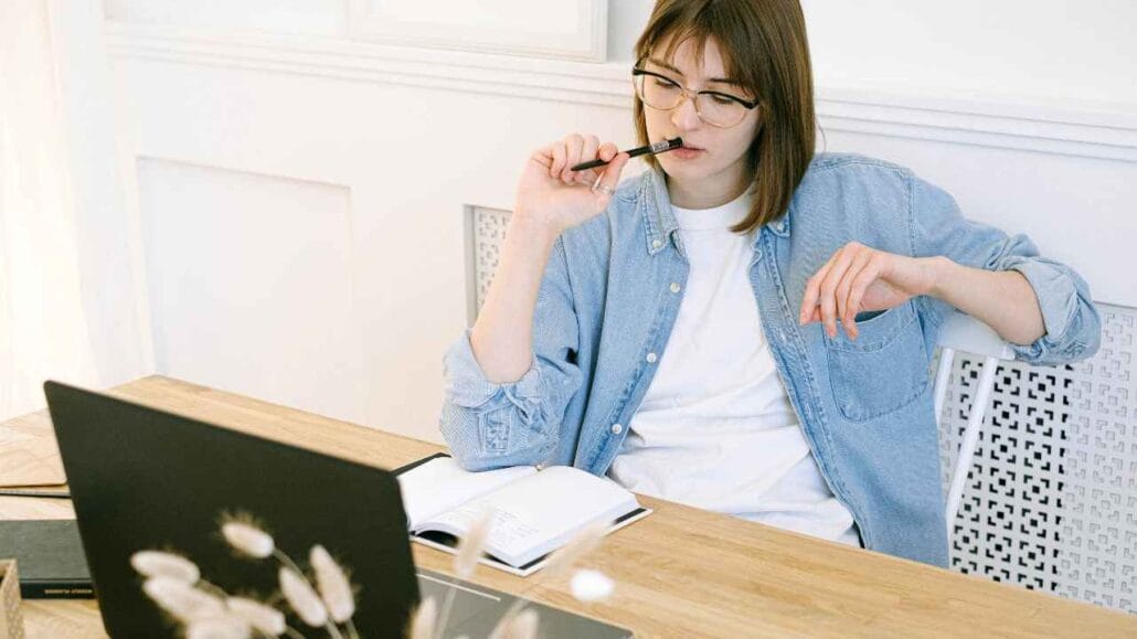 Mulher sentada à mesa com notebook aberto, segurando uma caneta próxima à boca enquanto analisa um caderno de anotações.