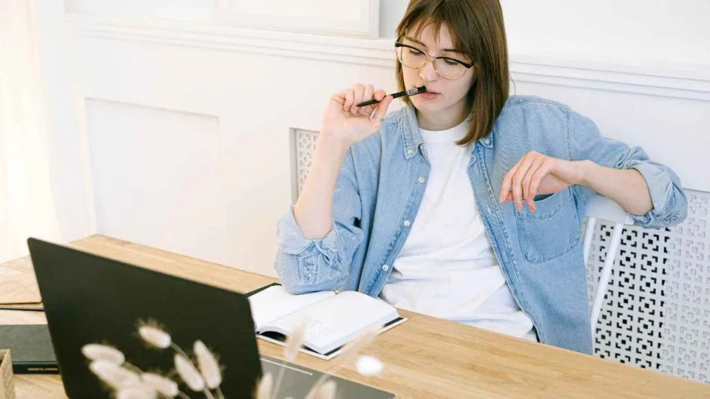 Mulher sentada à mesa com notebook aberto, segurando uma caneta próxima à boca enquanto analisa um caderno de anotações.