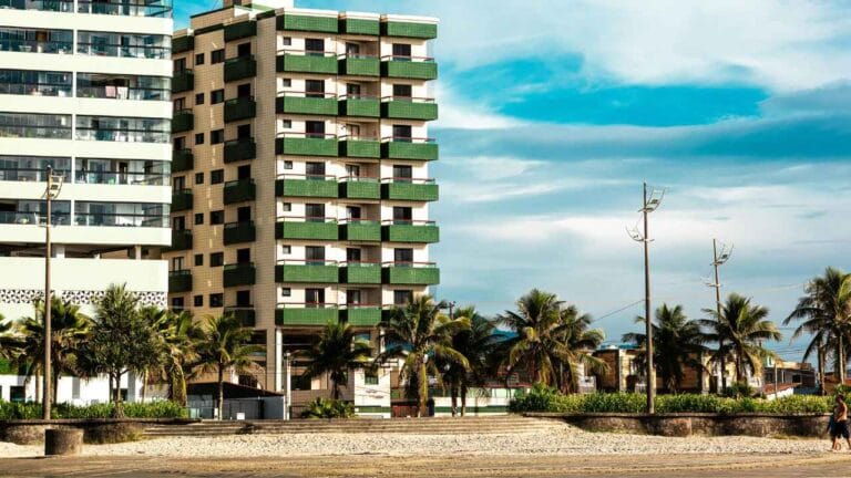 Prédio residencial à beira-mar com varandas verdes, palmeiras na calçada e céu azul ao fundo.