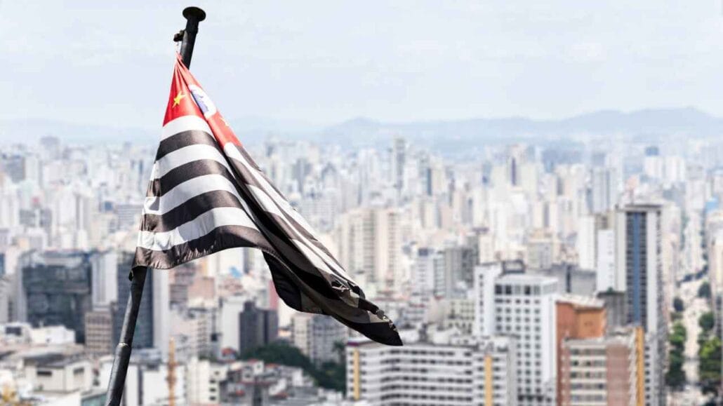 Bndeira de são paulo em destaque, atrás dela estão prédios.