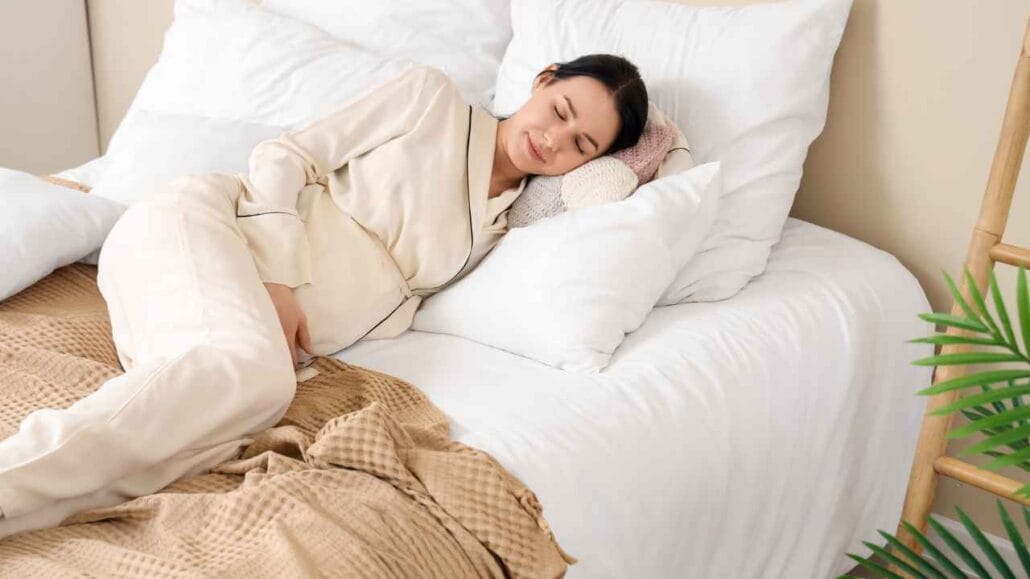 Mulher grávida dormindo de lado em cama com travesseiros brancos, vestindo pijama claro e apoiando a barriga.