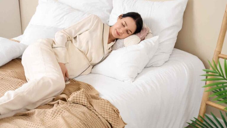 Mulher grávida dormindo de lado em cama com travesseiros brancos, vestindo pijama claro e apoiando a barriga.