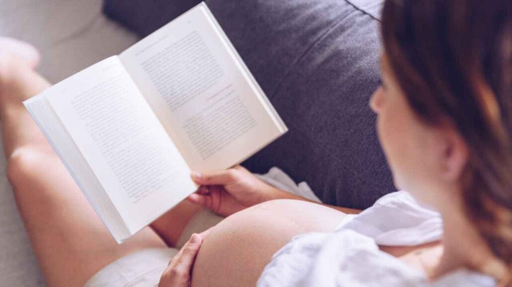 Mulher grávida sentada no sofá lendo um livro aberto apoiado nas mãos enquanto descansa.