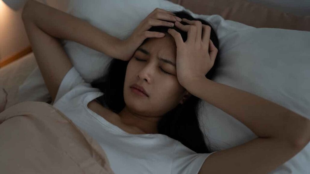 Mulher deitada na cama segurando a cabeça com as mãos, demonstrando desconforto com barulho para dormir
