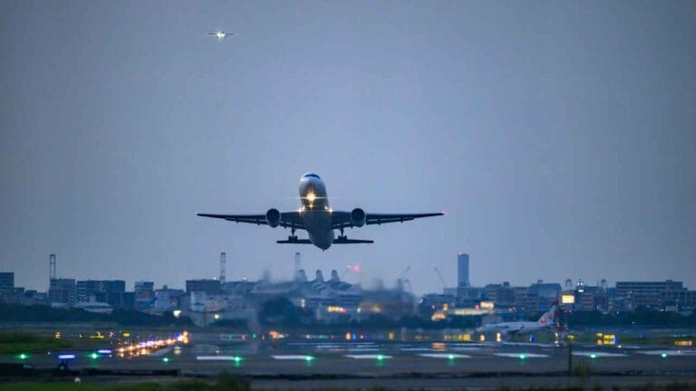 Avião em decolagem canva. Ao fundo uma cidade ao anoitecer.