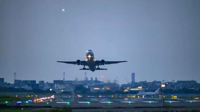 Avião em decolagem canva. Ao fundo uma cidade ao anoitecer.