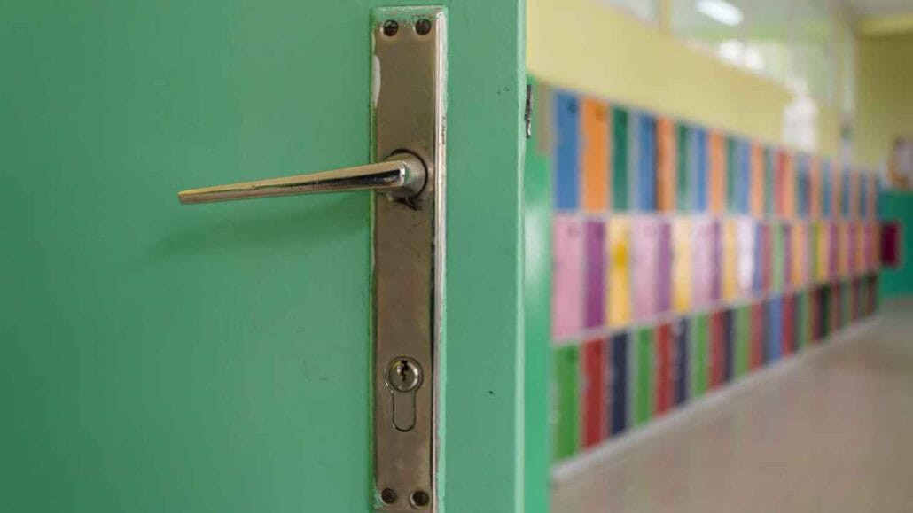 Porta verde parcialmente aberta com maçaneta metálica em destaque, revelando corredor escolar com armários coloridos ao fundo.