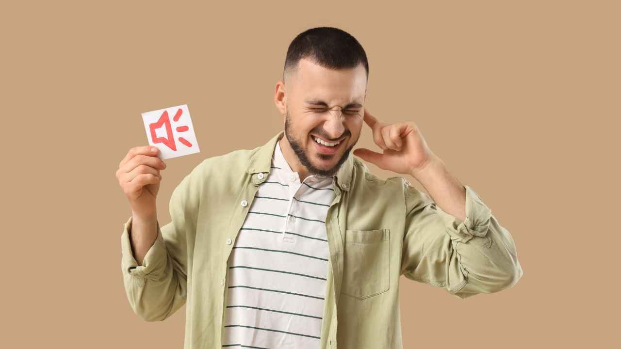 Homem tampa um dos ouvidos enquanto faz expressão de desconforto e segura um cartão com símbolo de alto-falante, representando incômodo causado decibeis ou vibrações sonoras