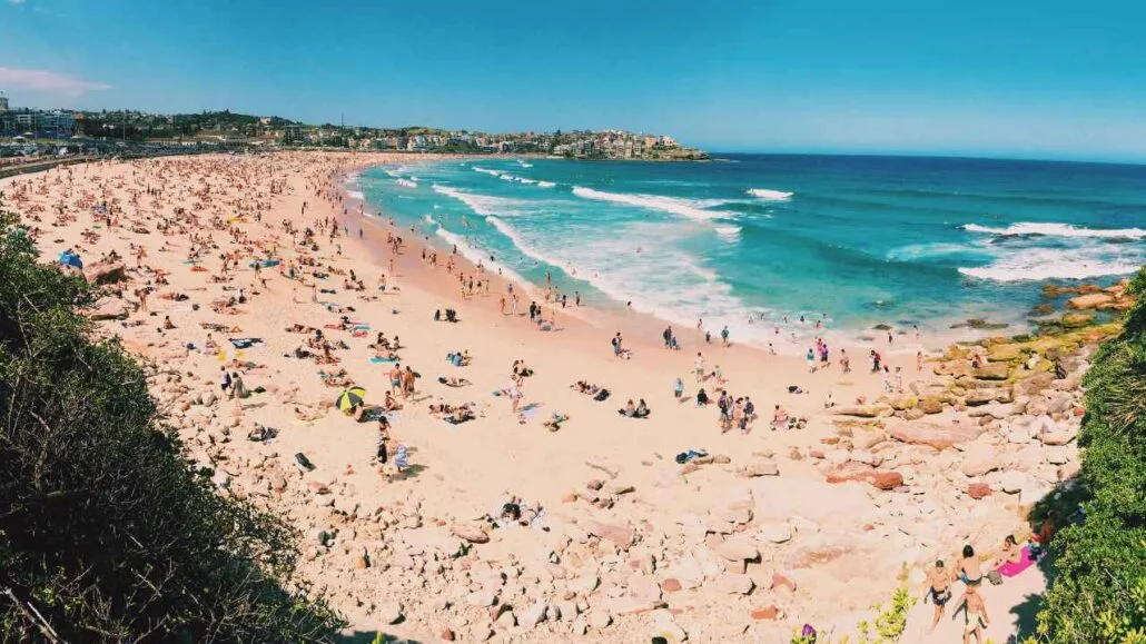 Praia movimentada em dia ensolarado, com grande número de pessoas na areia e no mar