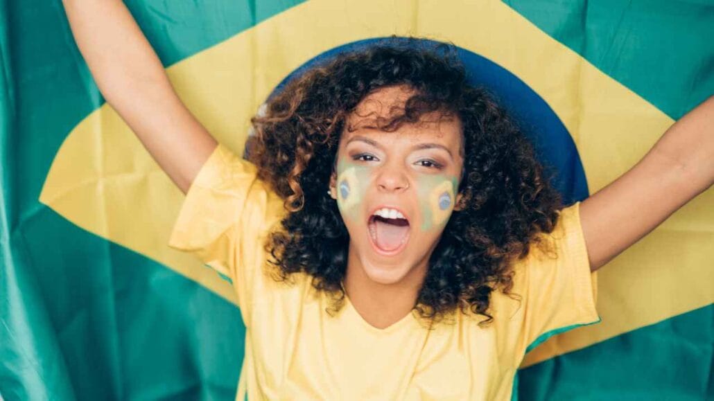 Jovem torcedora com pintura verde e amarela no rosto comemora com os braços erguidos em frente à bandeira do Brasil